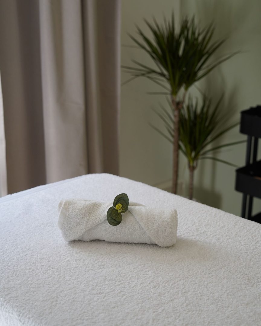 A bright treatment room with a rolled white towel on a linen-covered bed beside a potted palm